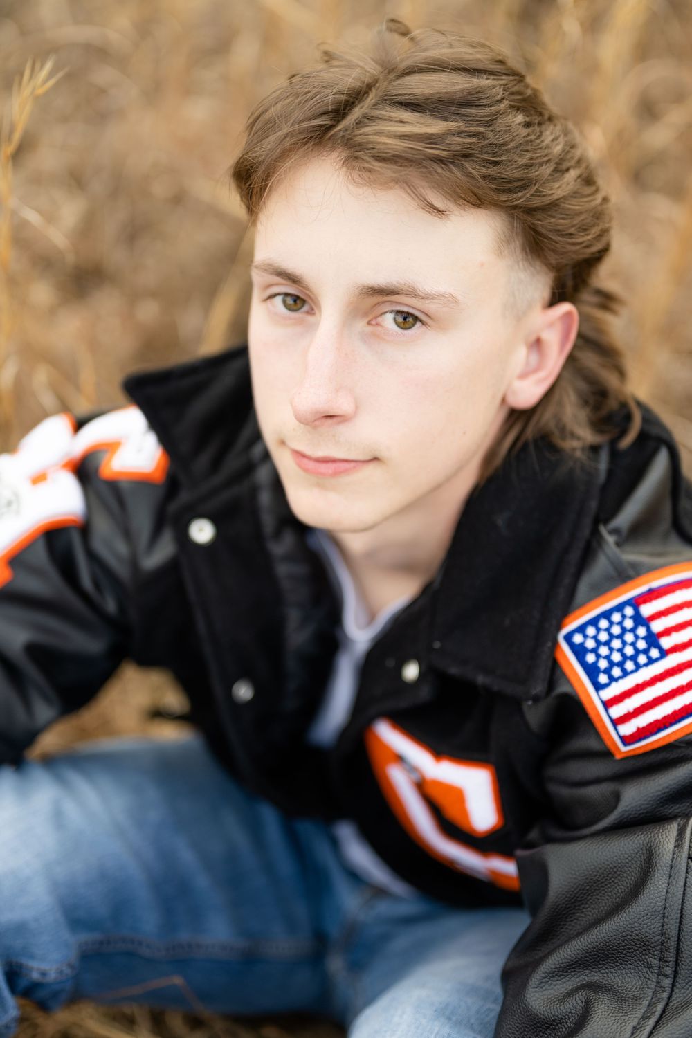 Portrait of a person in a black jacket with American flag patches sitting in tall grass.