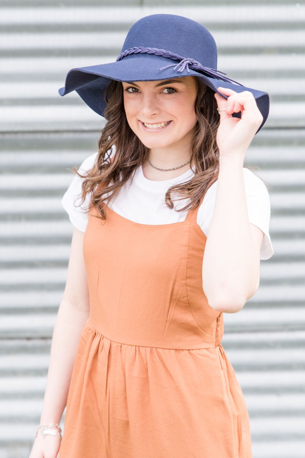 A person in an orange dress and navy hat smiles while standing against a striped wall.