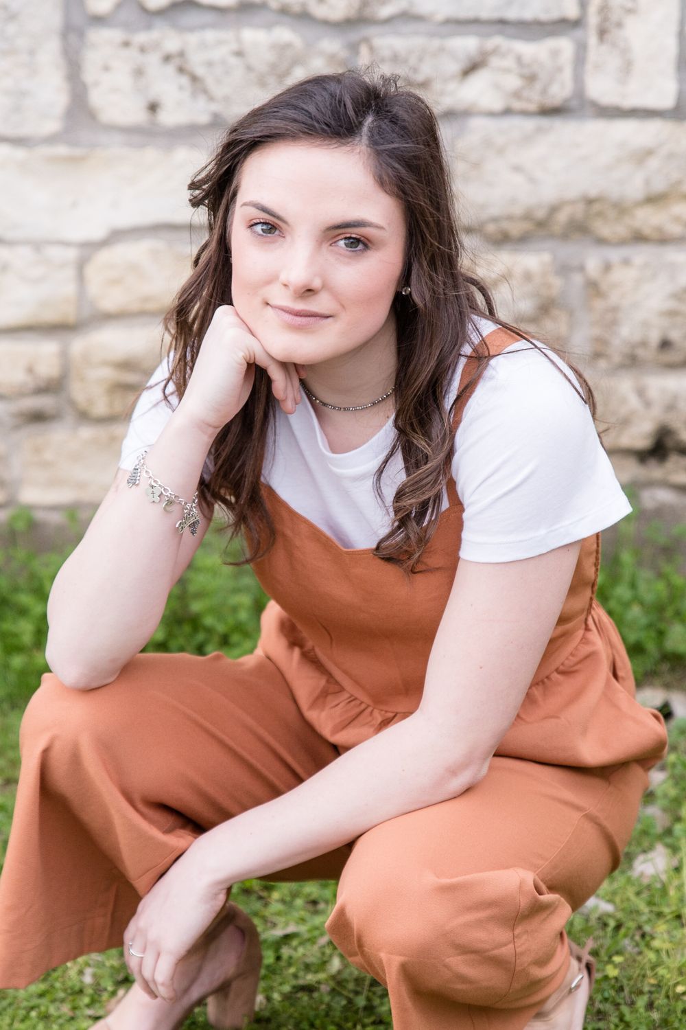 A casual portrait of someone wearing an orange overall dress against a stone wall background.