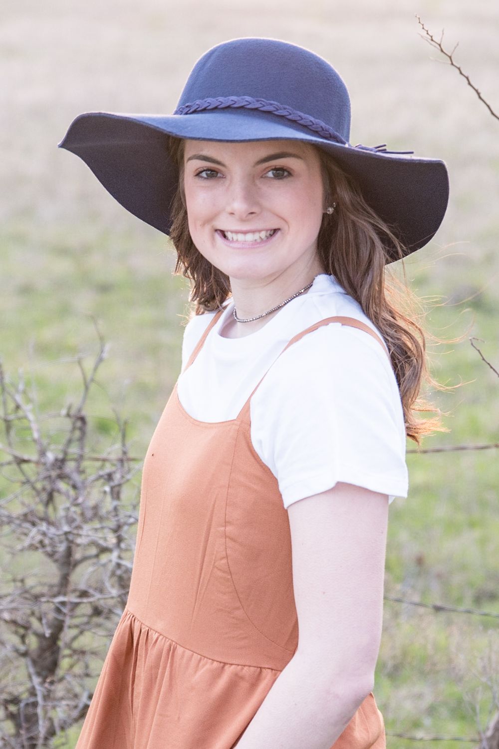 A woman in an orange jumper dress and navy wide-brim hat poses in an outdoor setting.