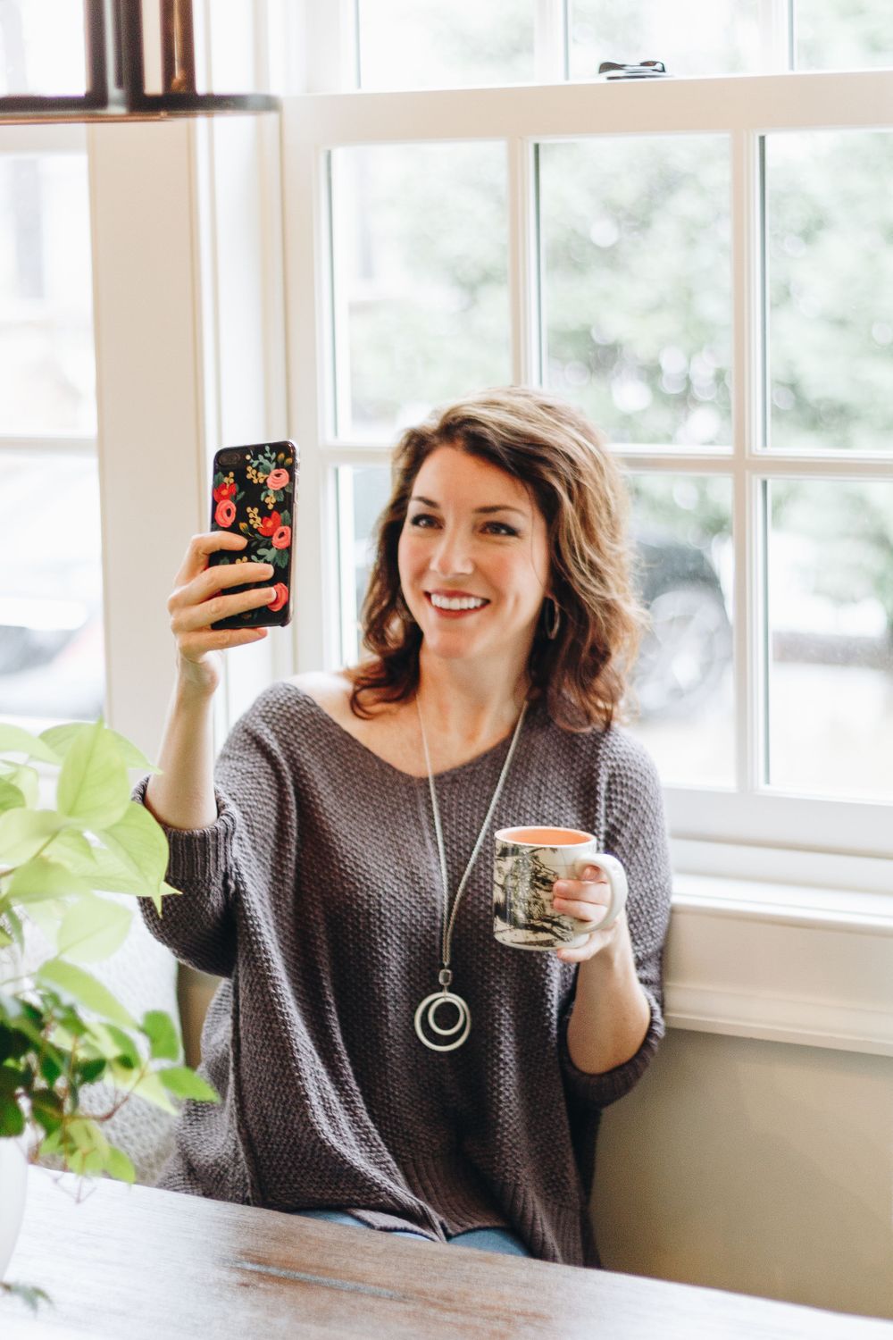 Taking a selfie by a bright window while holding coffee and wearing a cozy gray sweater.