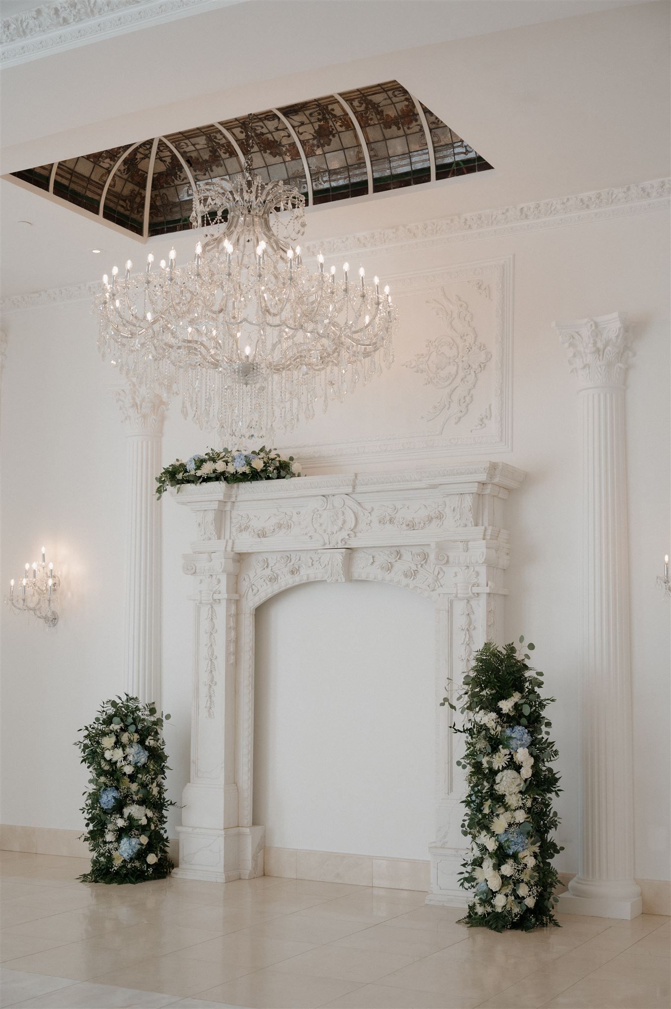 Elegant white wedding altar with ornate fireplace mantel, crystal chandelier, and floral arrangements in a luxurious room.