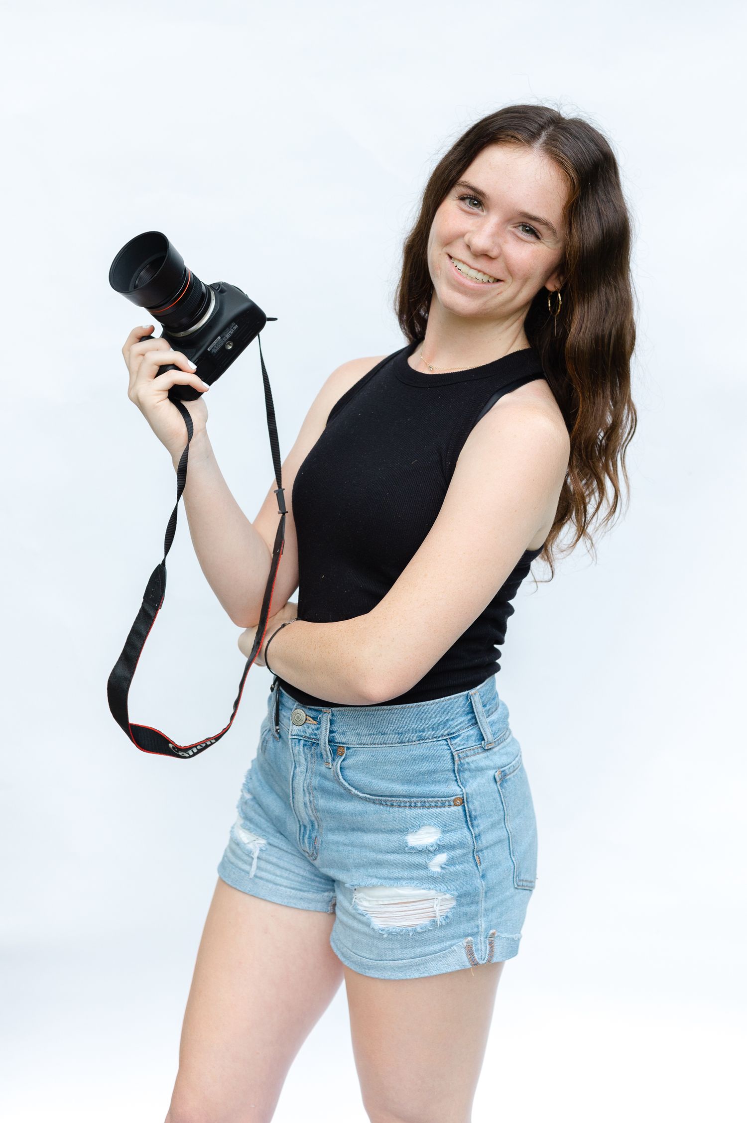 Photographer wearing black sleeveless top and denim shorts holds professional camera against white background.