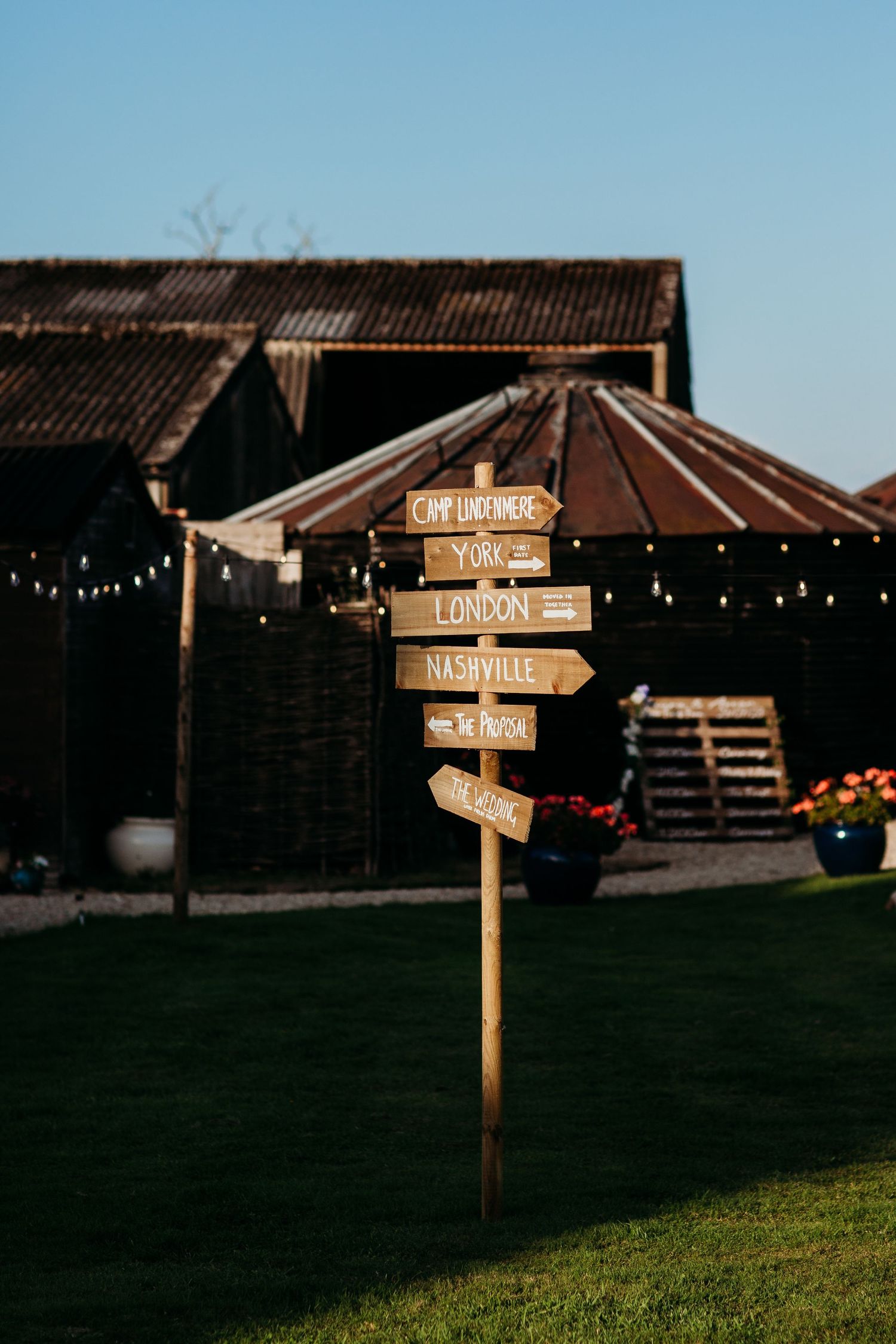 A rustic wooden directional sign post with hanging string lights at an outdoor wedding venue.