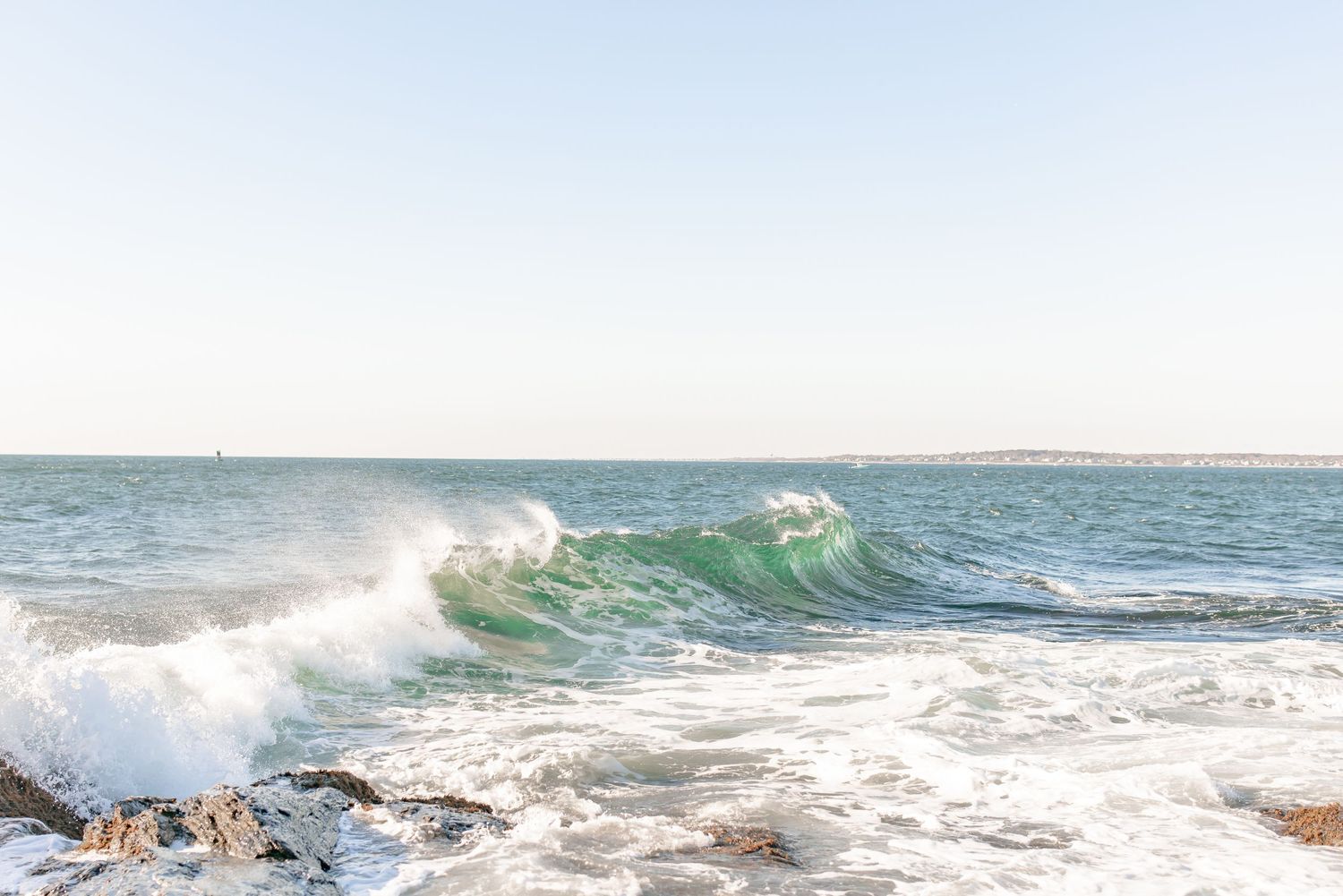 Vibrant green ocean wave crashing along a rocky shoreline under a clear blue sky.