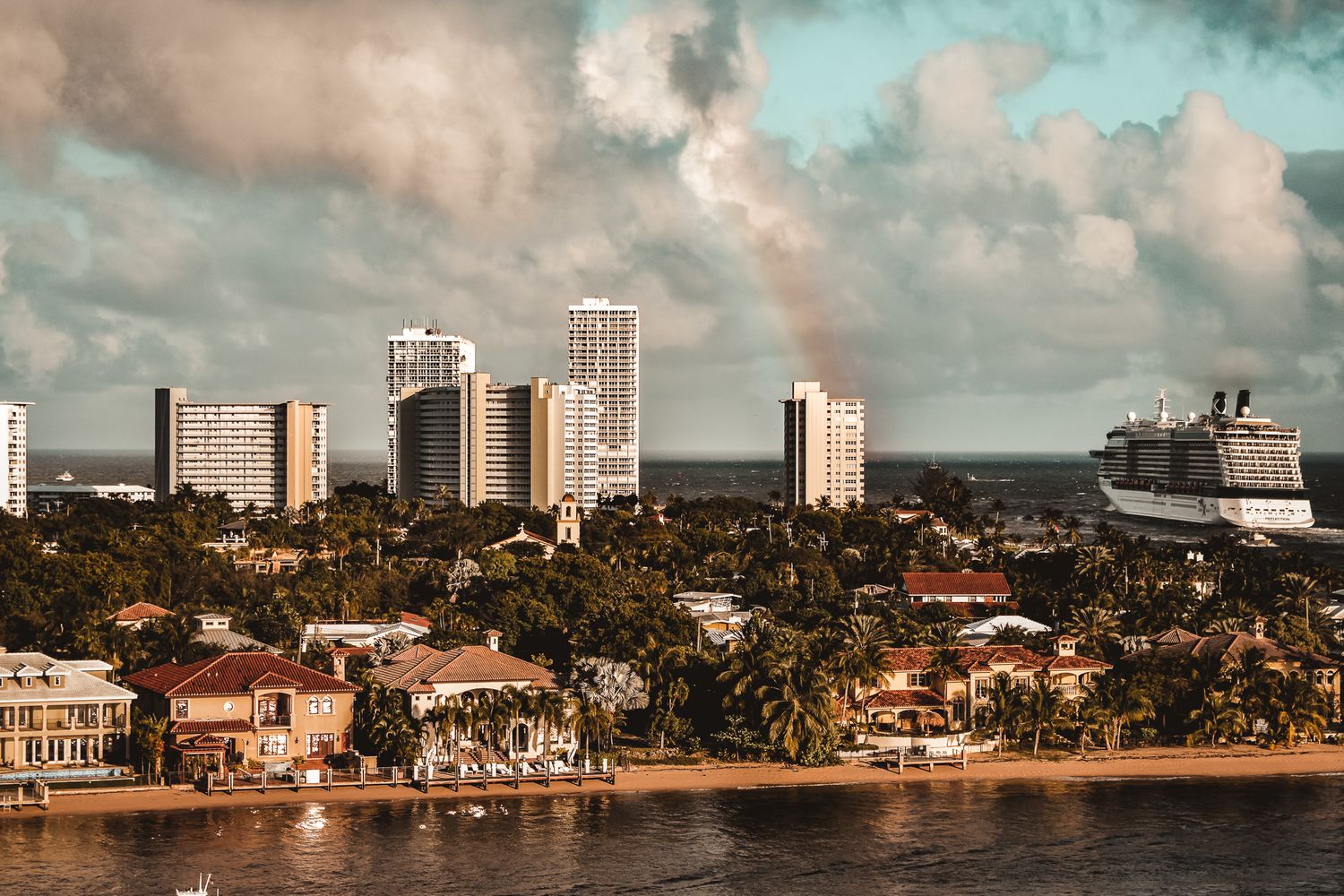 Coastal city skyline with high-rise buildings and cruise ship visible in warm sunset light.