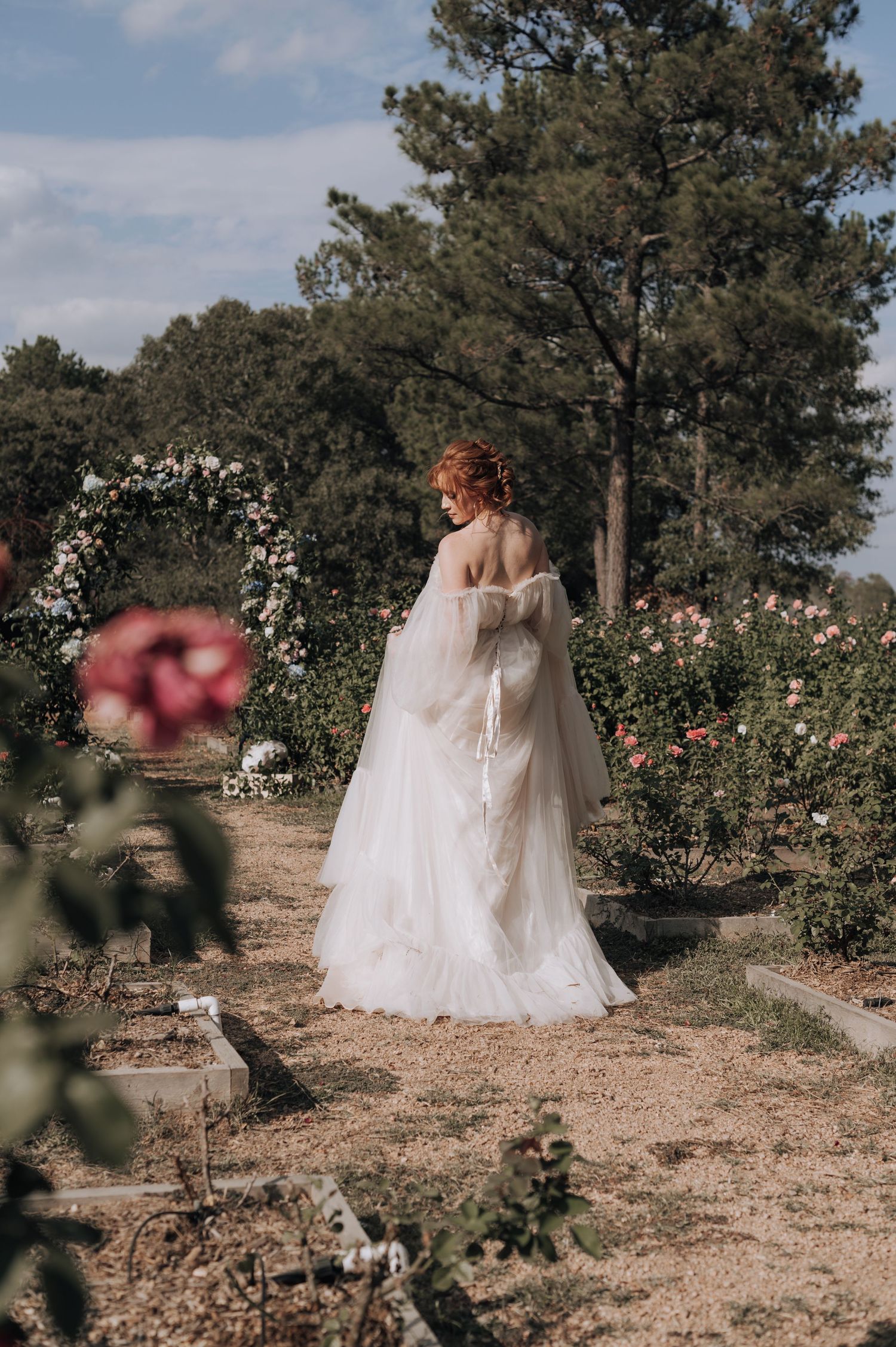 Classic wedding gown silhouette against floral arch and pine trees in garden setting.