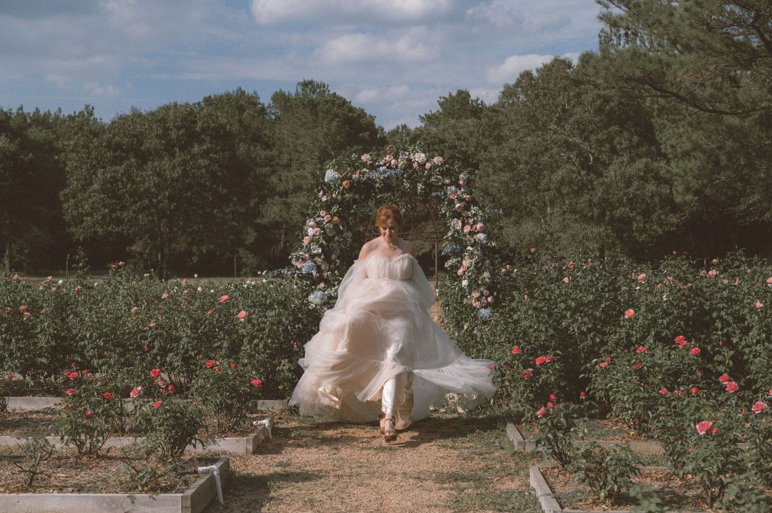 Ethereal wedding dress flowing in a romantic garden setting with a floral arch and blooming roses.