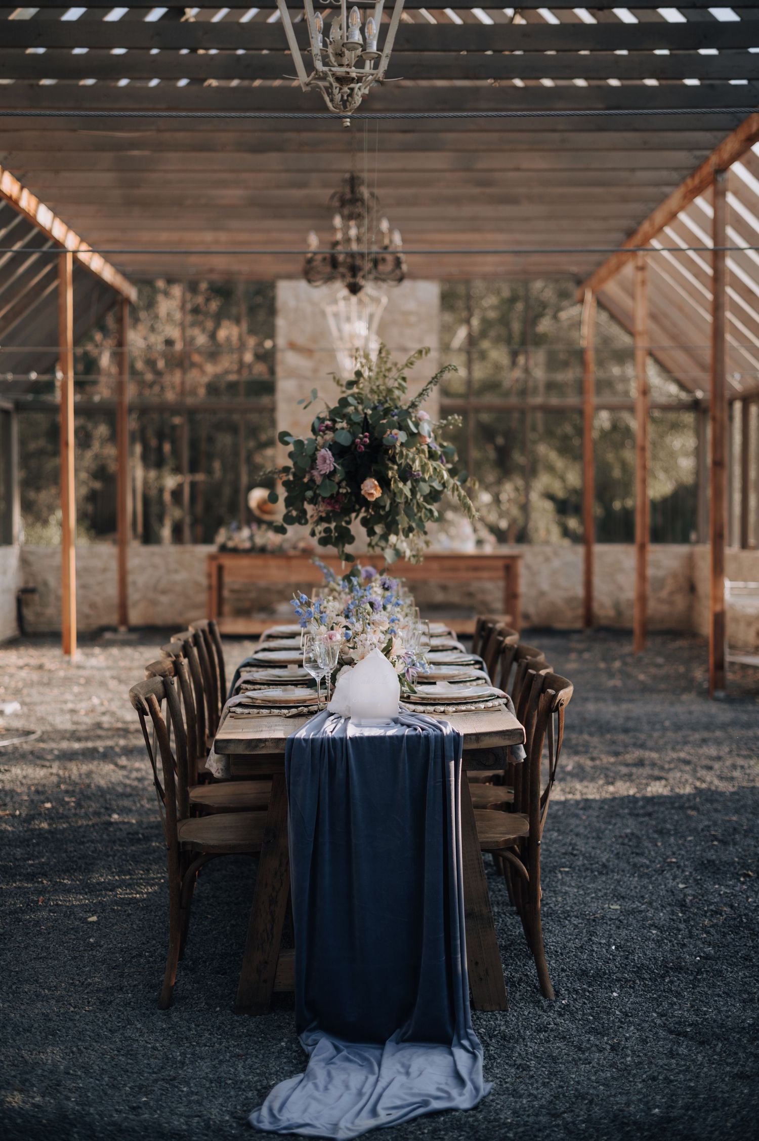 Elegant wedding reception table with navy runner and wooden chairs under a covered rustic outdoor venue with chandeliers.
