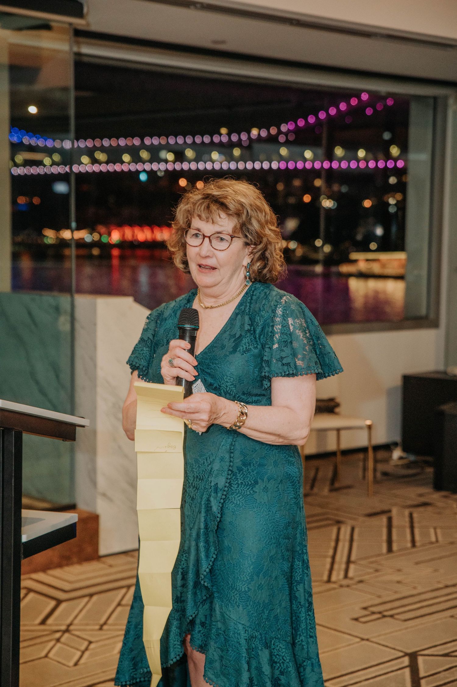 Woman in teal dress gives speech at evening reception with illuminated bridge visible through windows.