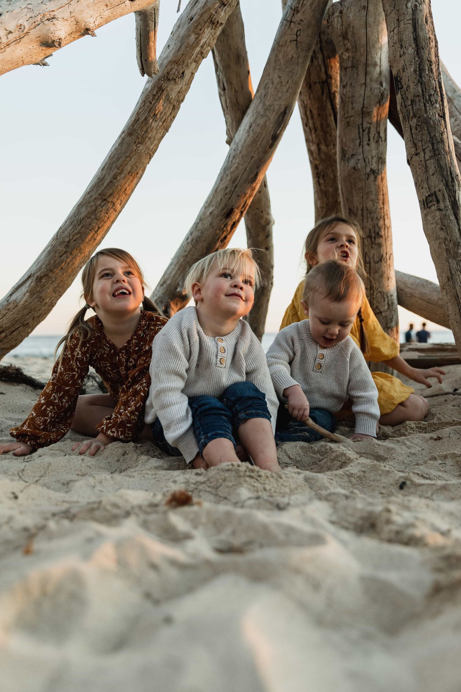 Four young children sit together in sand beneath a driftwood structure at the beach.