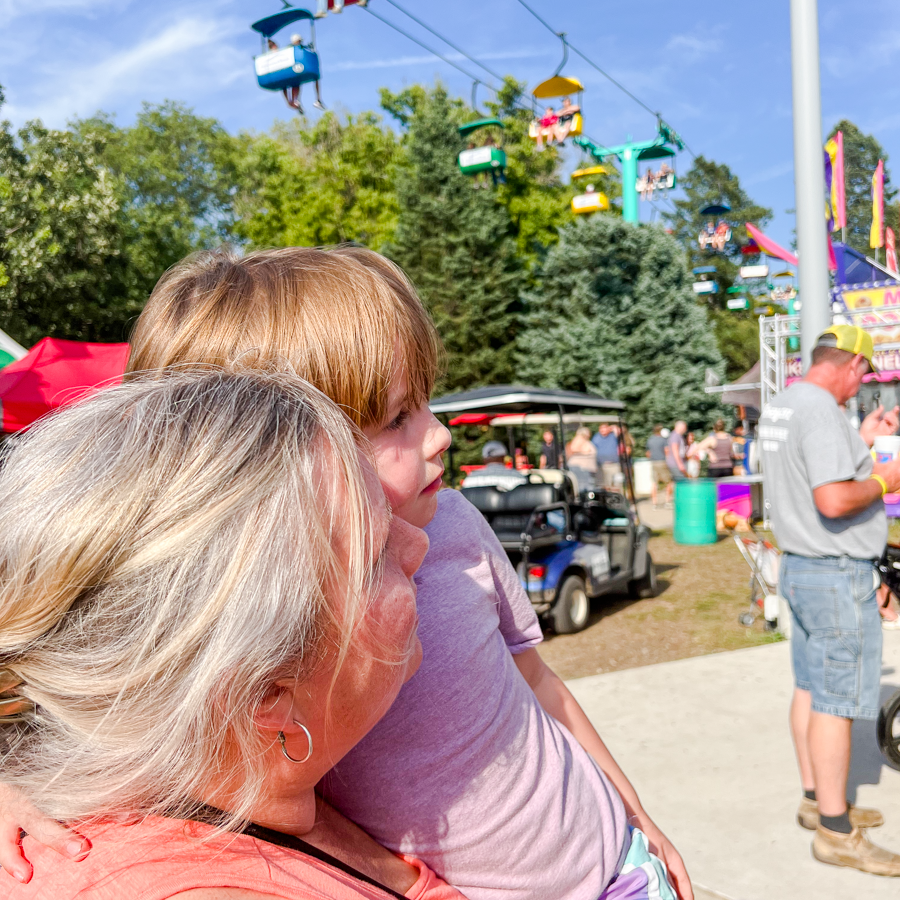 Visitors watching colorful overhead ski lift chairs moving across a sunny fairground.