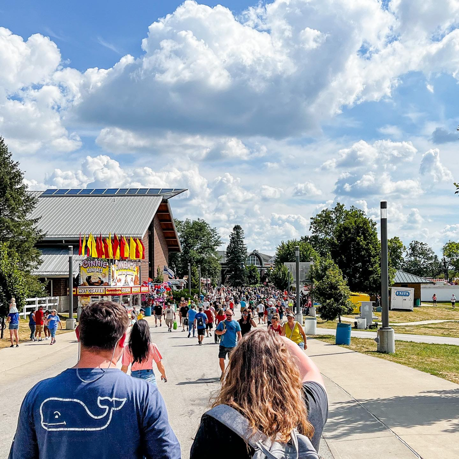 Crowded fairground walkway with colorful vendor stands and fluffy white clouds overhead.