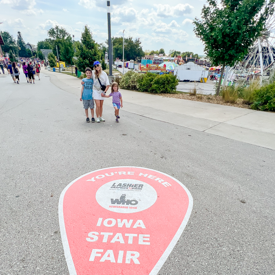 Pink Iowa State Fair directional marker on pavement with fairgoers walking in the background.