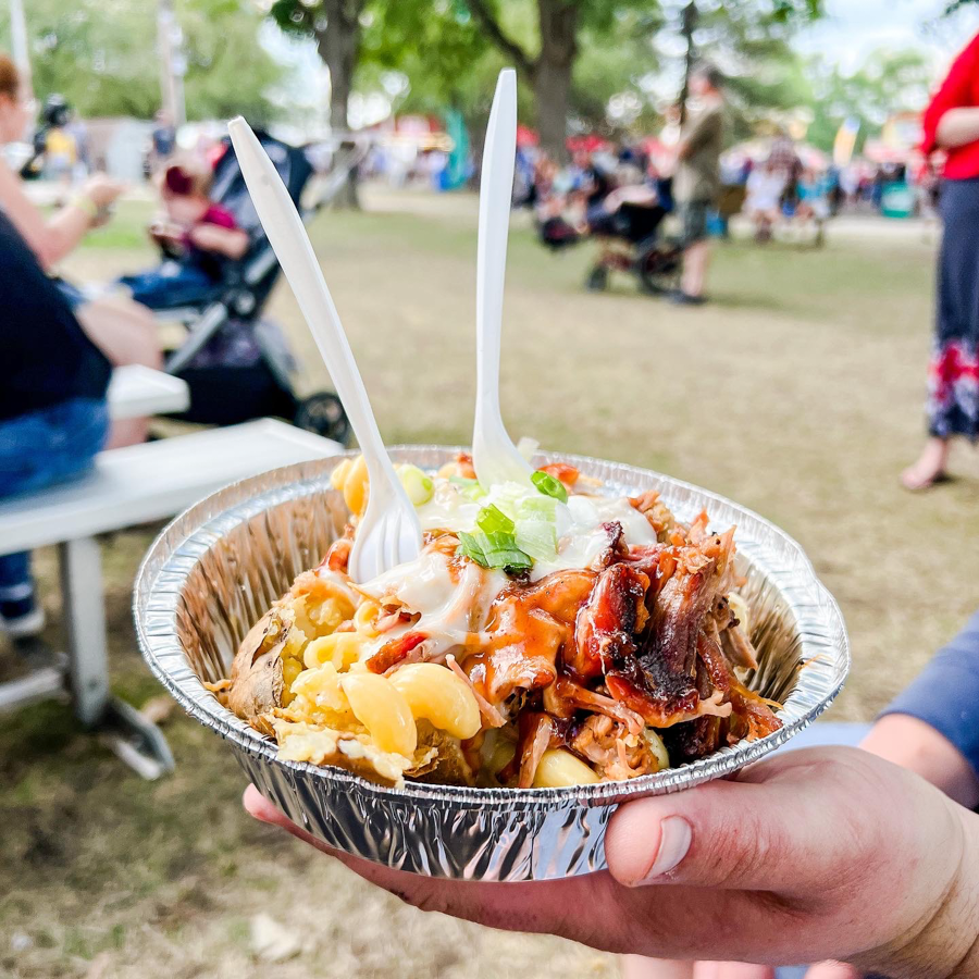 Loaded nachos in aluminum container with toppings and plastic forks at an outdoor event.