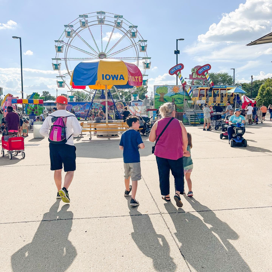 Panoramic view of a crowded state fair midway with a ferris wheel visible in the distance on a sunny day.
