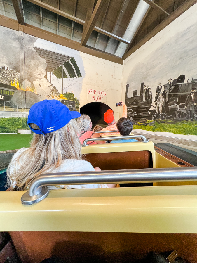 Riders on a vintage train ride passing by historical murals depicting railroad scenes.