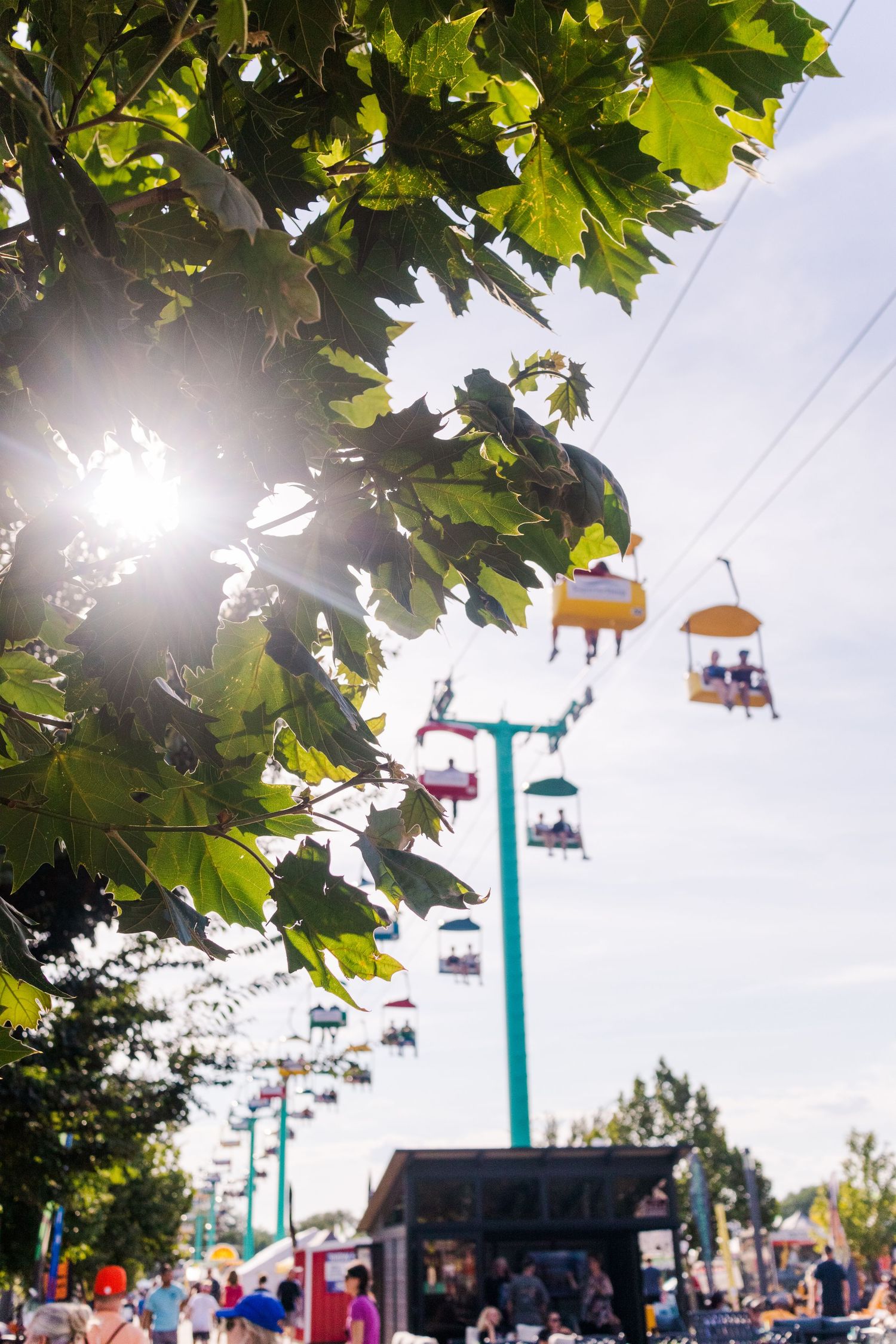 Overhead carnival skyride with colorful passenger cars framed by sunlit tree leaves.