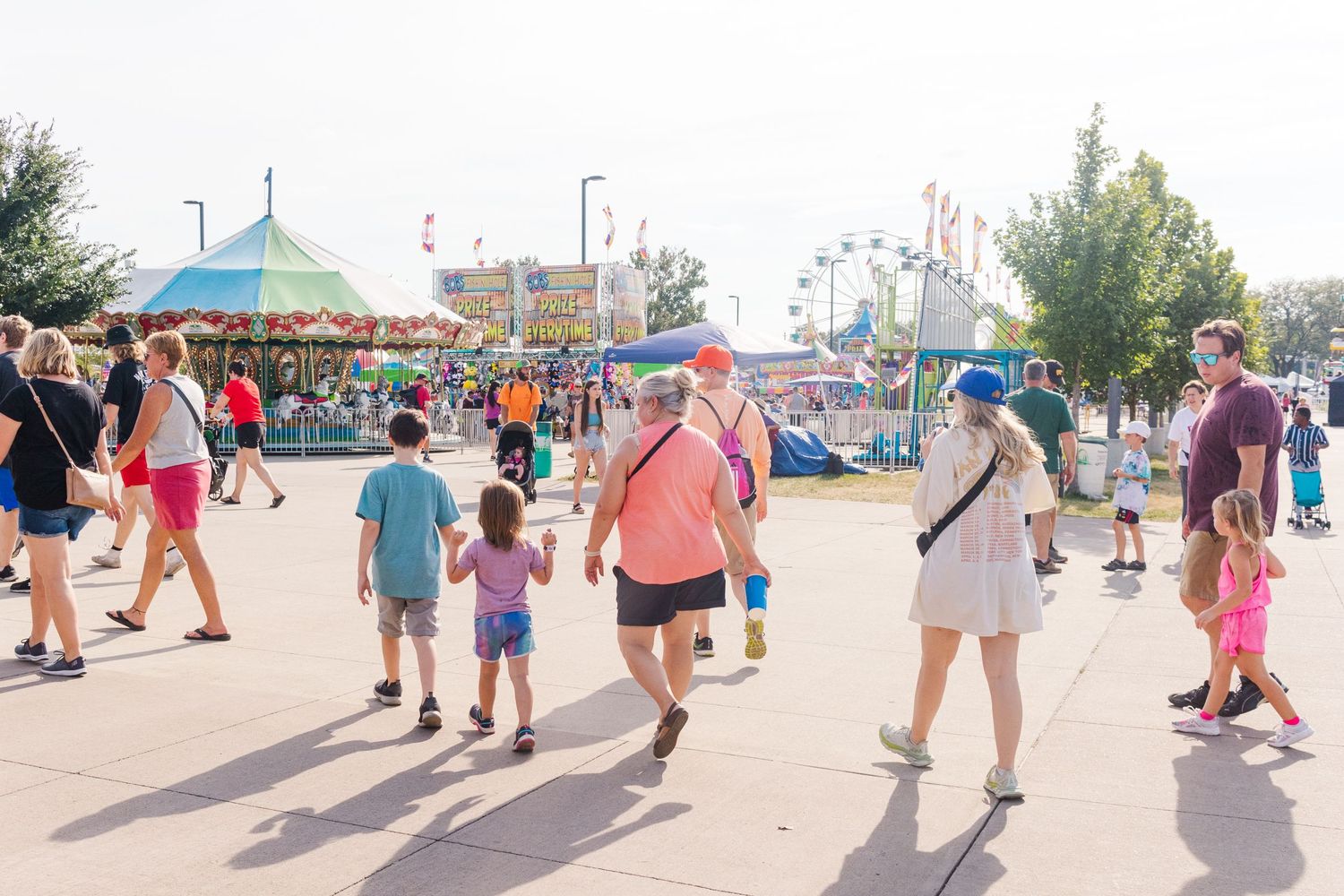 People walking through a sunny carnival midway with rides in the background.
