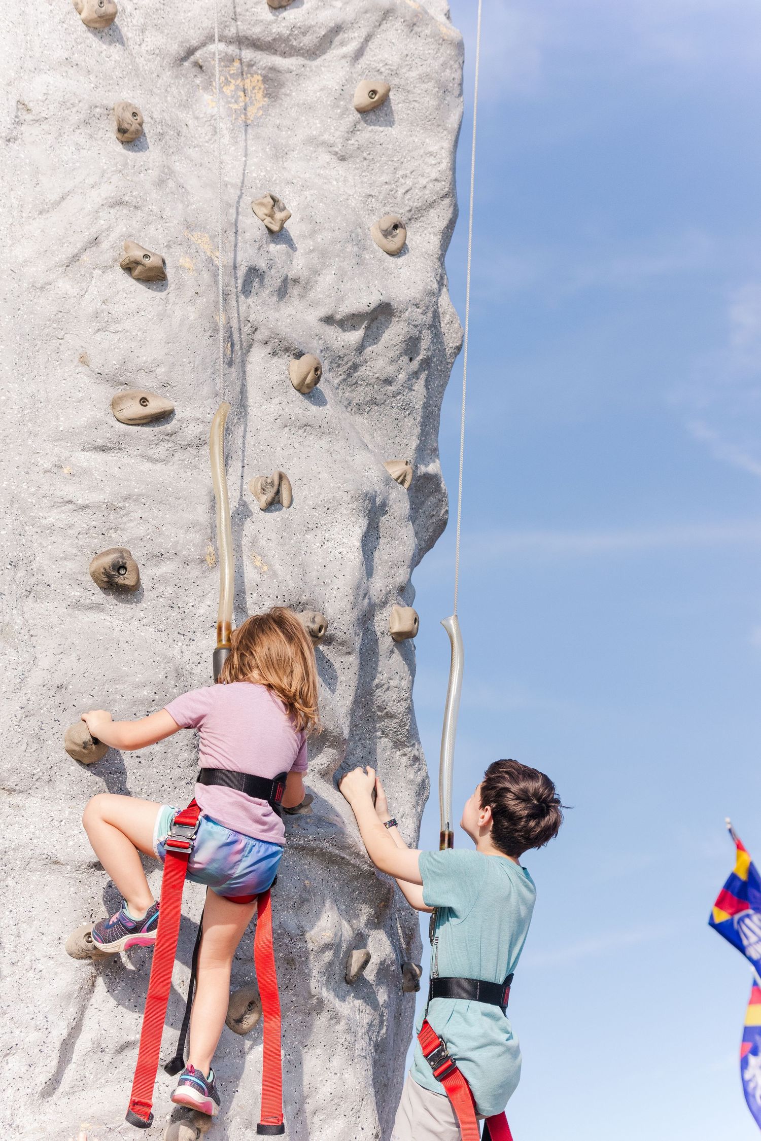 Two climbers scaling an outdoor climbing wall with safety harnesses.