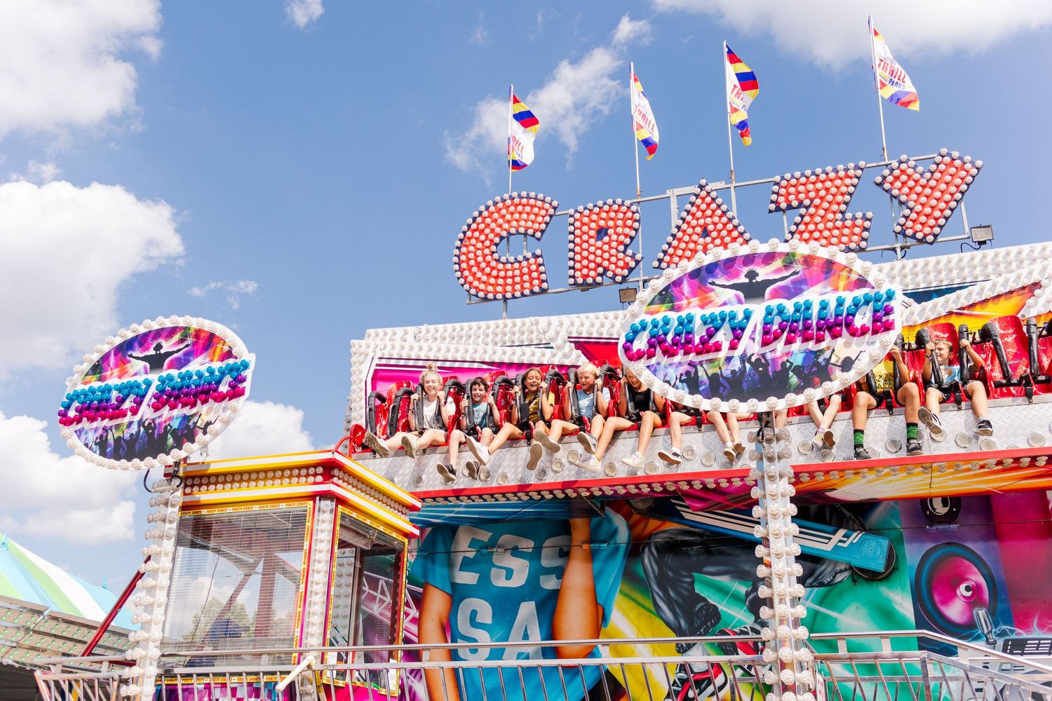 Bright carnival rides and game stalls with neon signage against a blue summer sky.