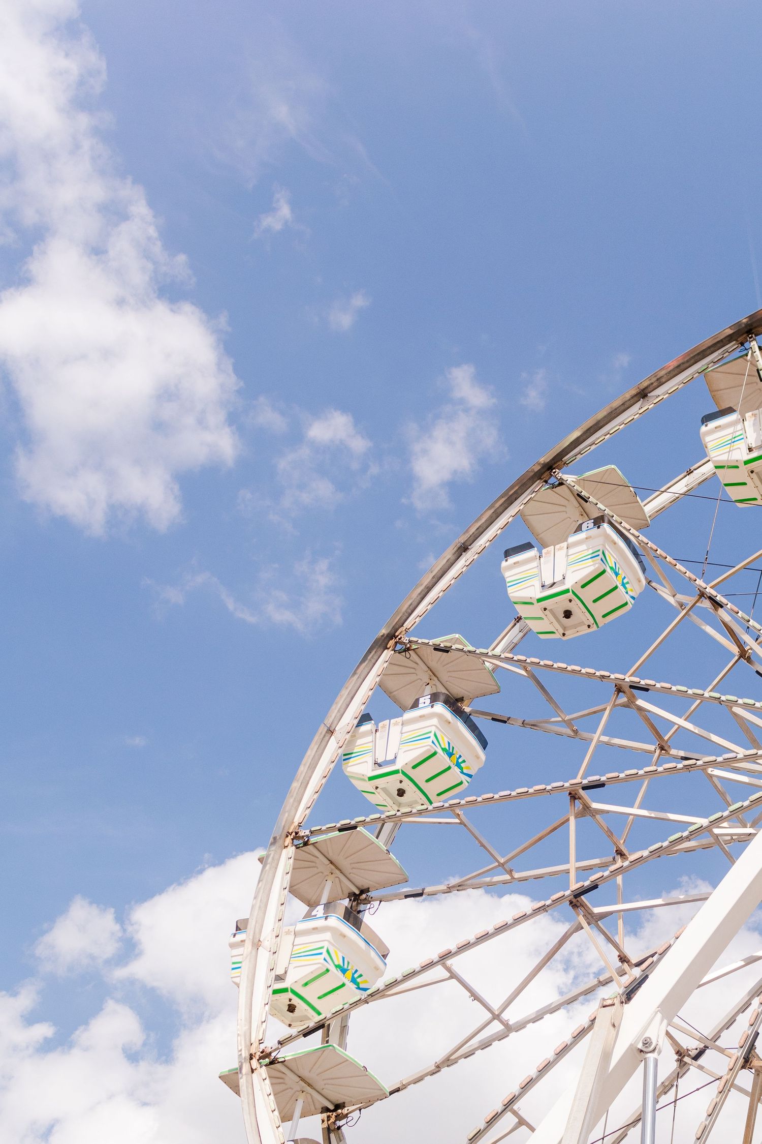 White metal framework of a Ferris wheel against a bright blue sky with puffy clouds.
