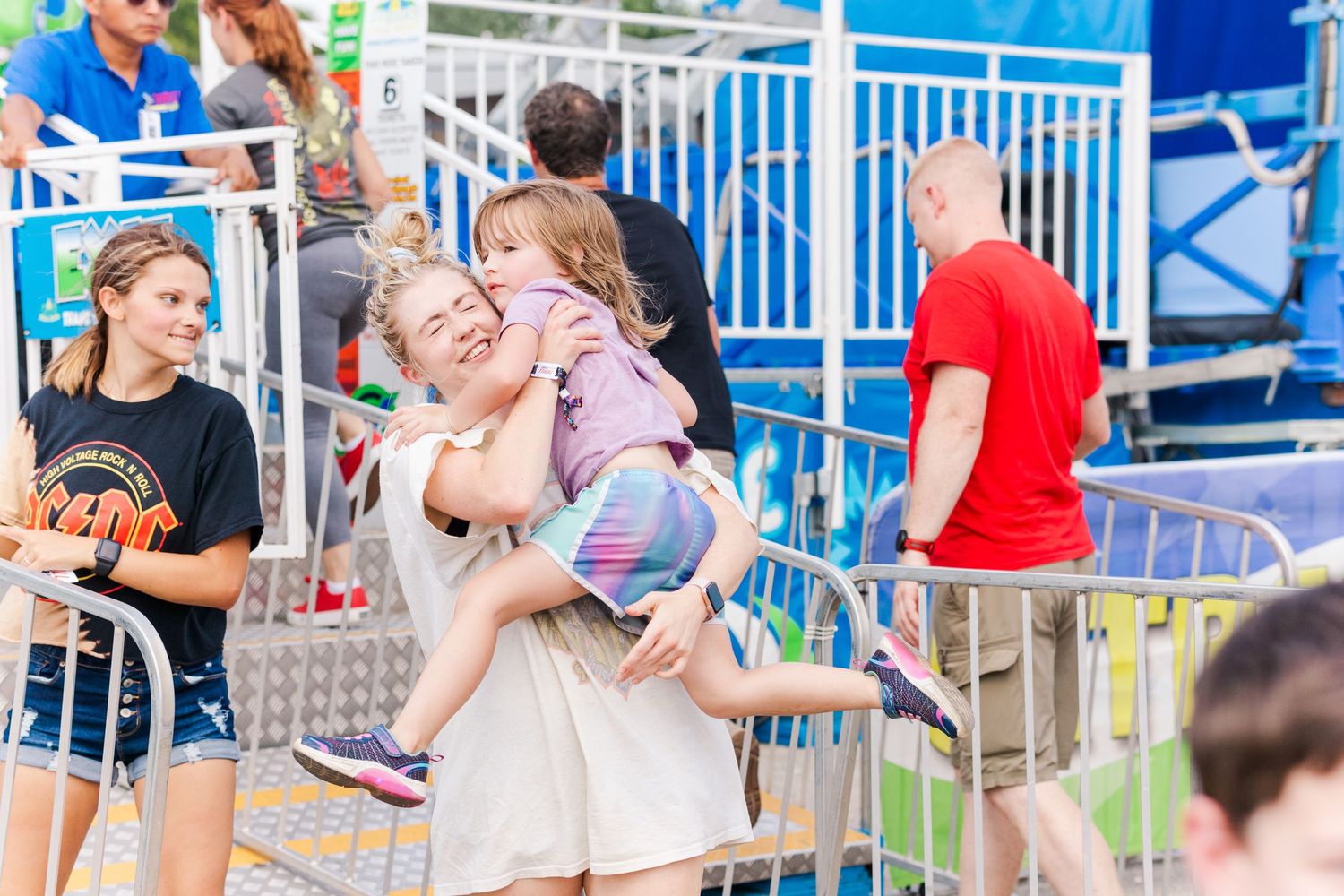 Fairground visitors walking past blue metal railings and carnival attractions.