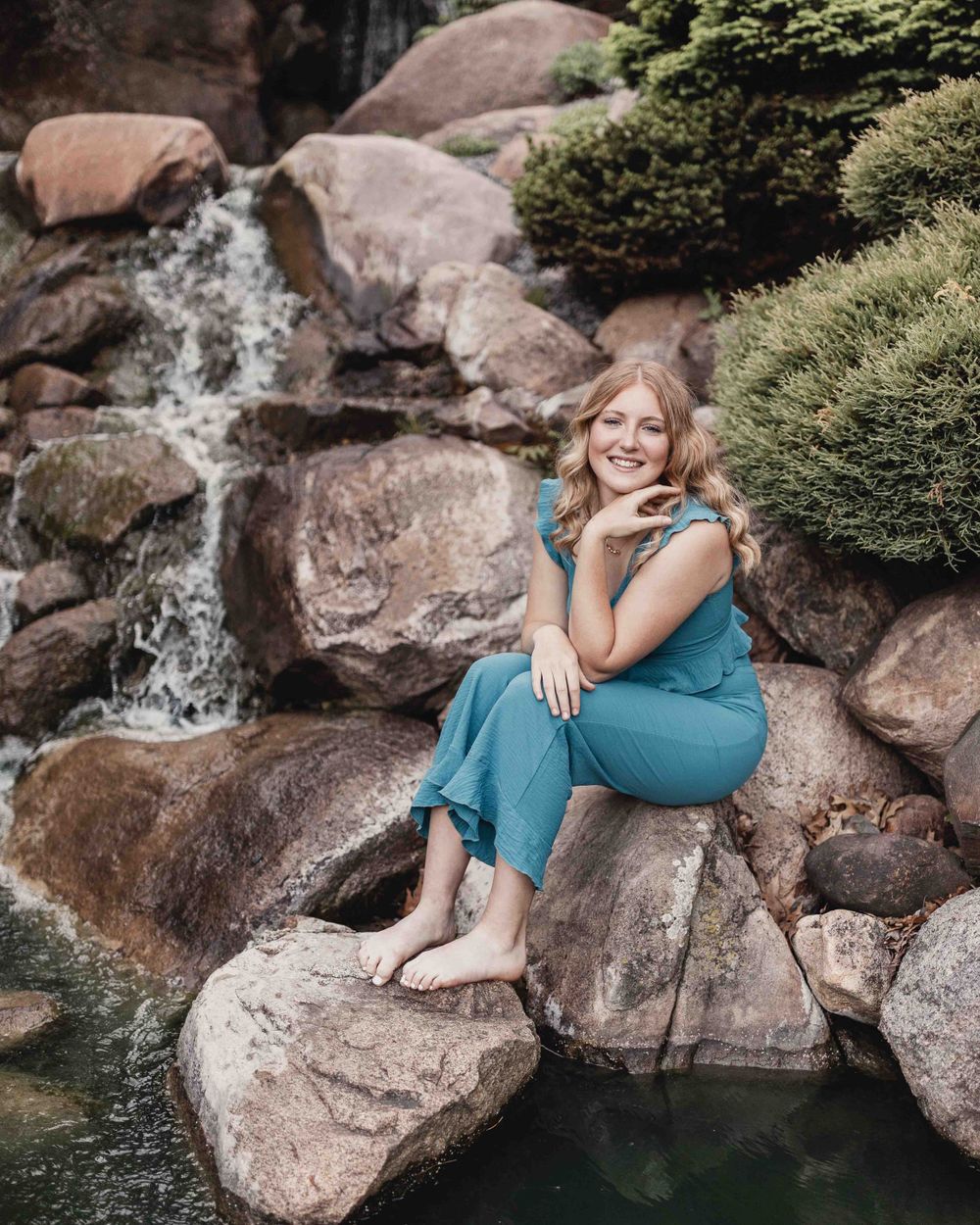 Senior portrait of female sitting by waterfall smiling outdoors at the Dubuque Iowa Arboretum and Botanical Gardens