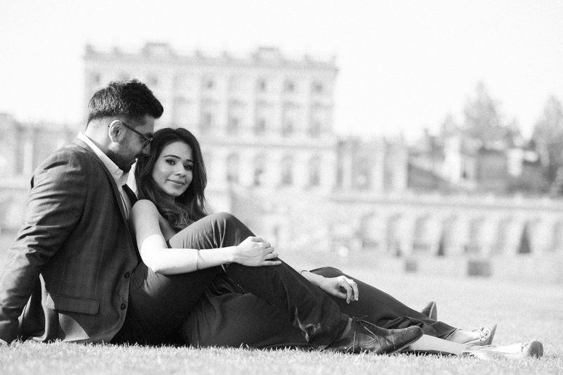 A romantic black and white photo of a couple relaxing together on a lawn with an elegant building in the background.