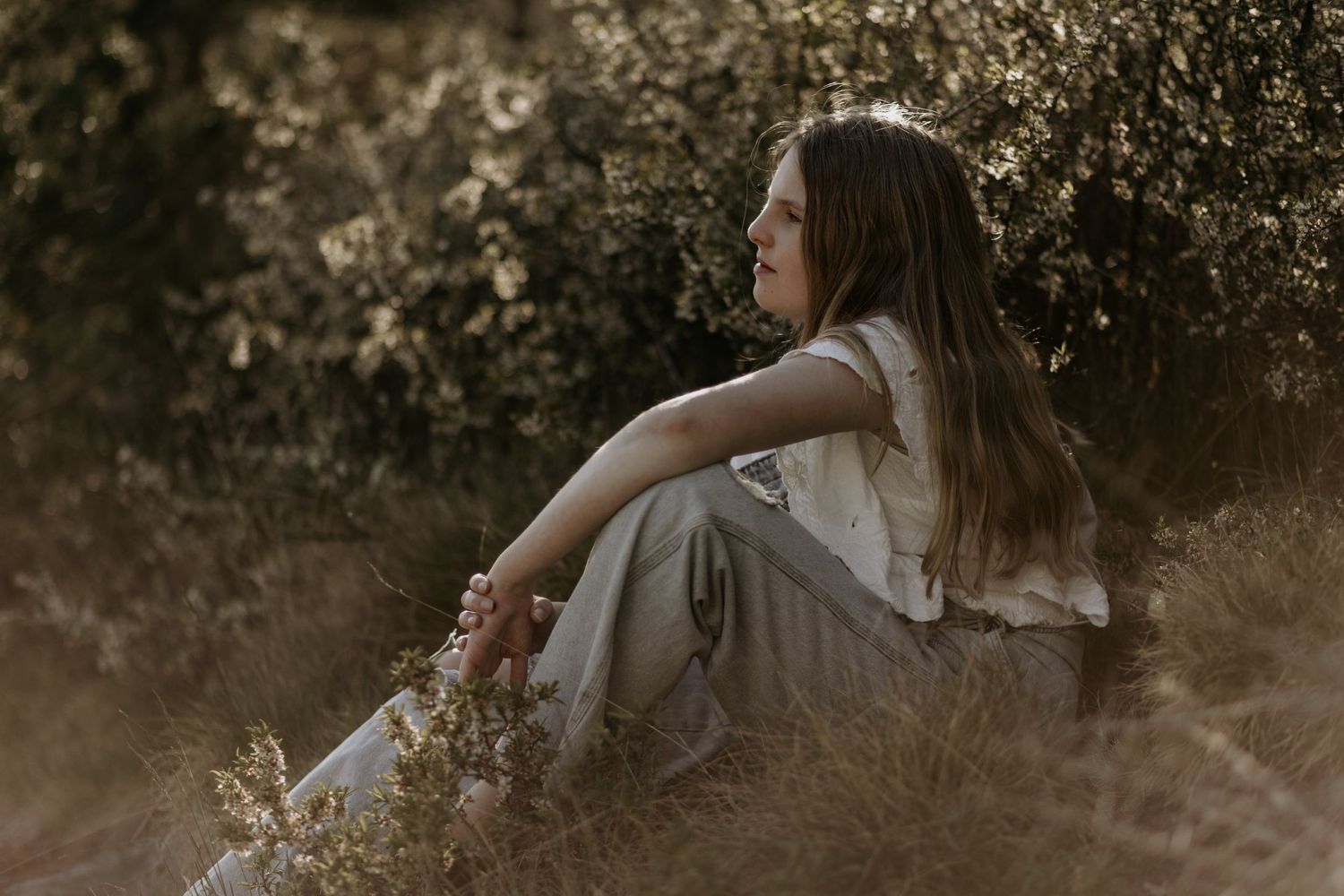 Person in casual clothes sitting thoughtfully in tall grass during golden hour.