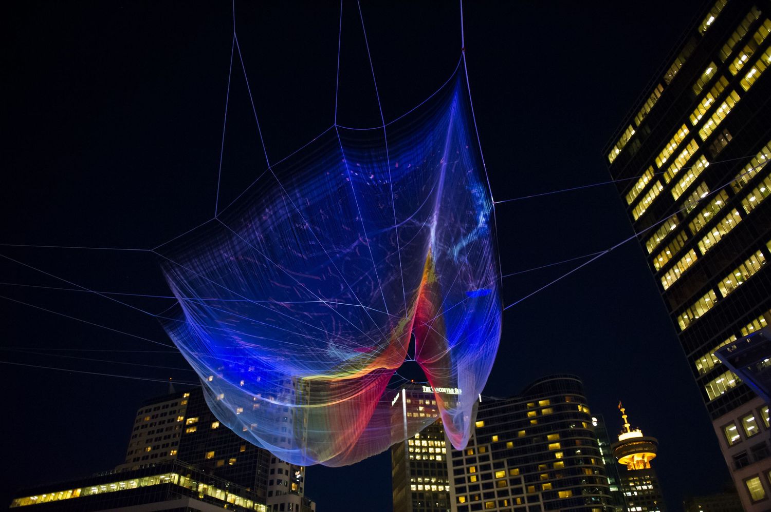 Large-scale illuminated fiber art installation glows blue and red against night sky in urban setting.