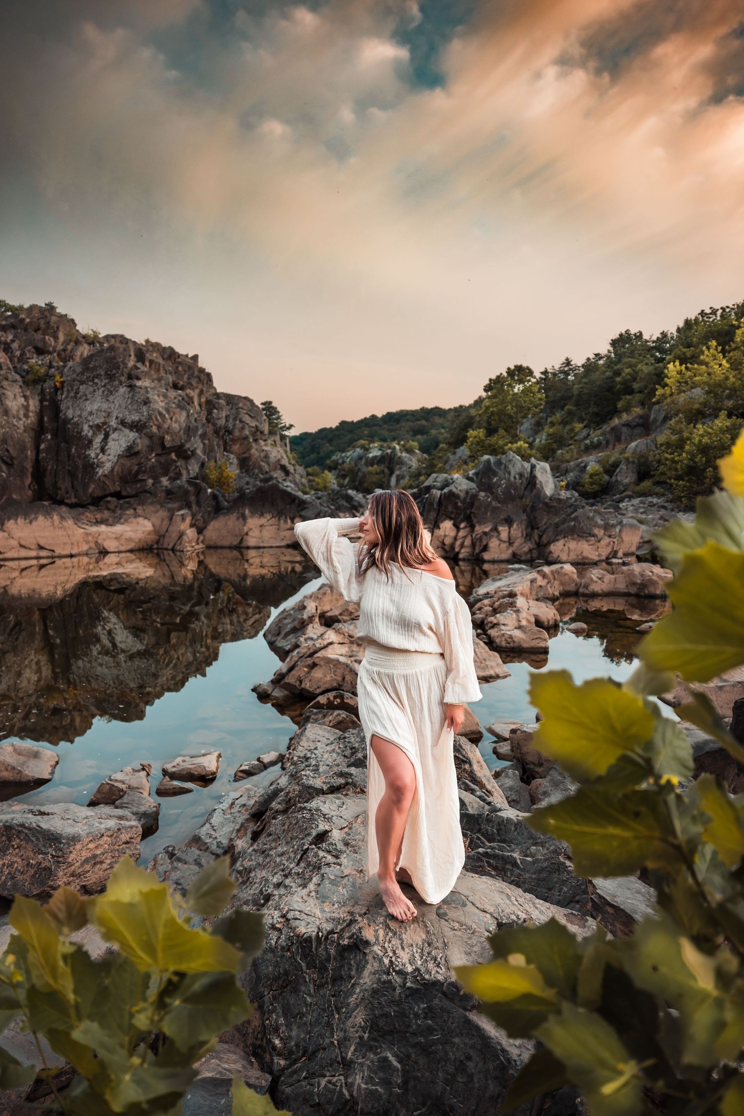 Woman in white dress poses on rocky terrain with dramatic sky and water reflections.