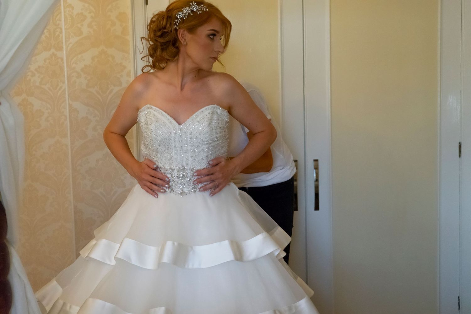 Bride in layered white wedding dress with ruffled skirt poses against cream-colored wall.