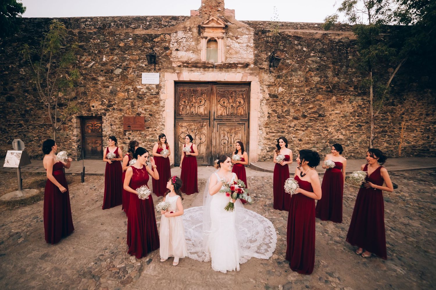 A bridal party in burgundy dresses poses outside a rustic stone church.