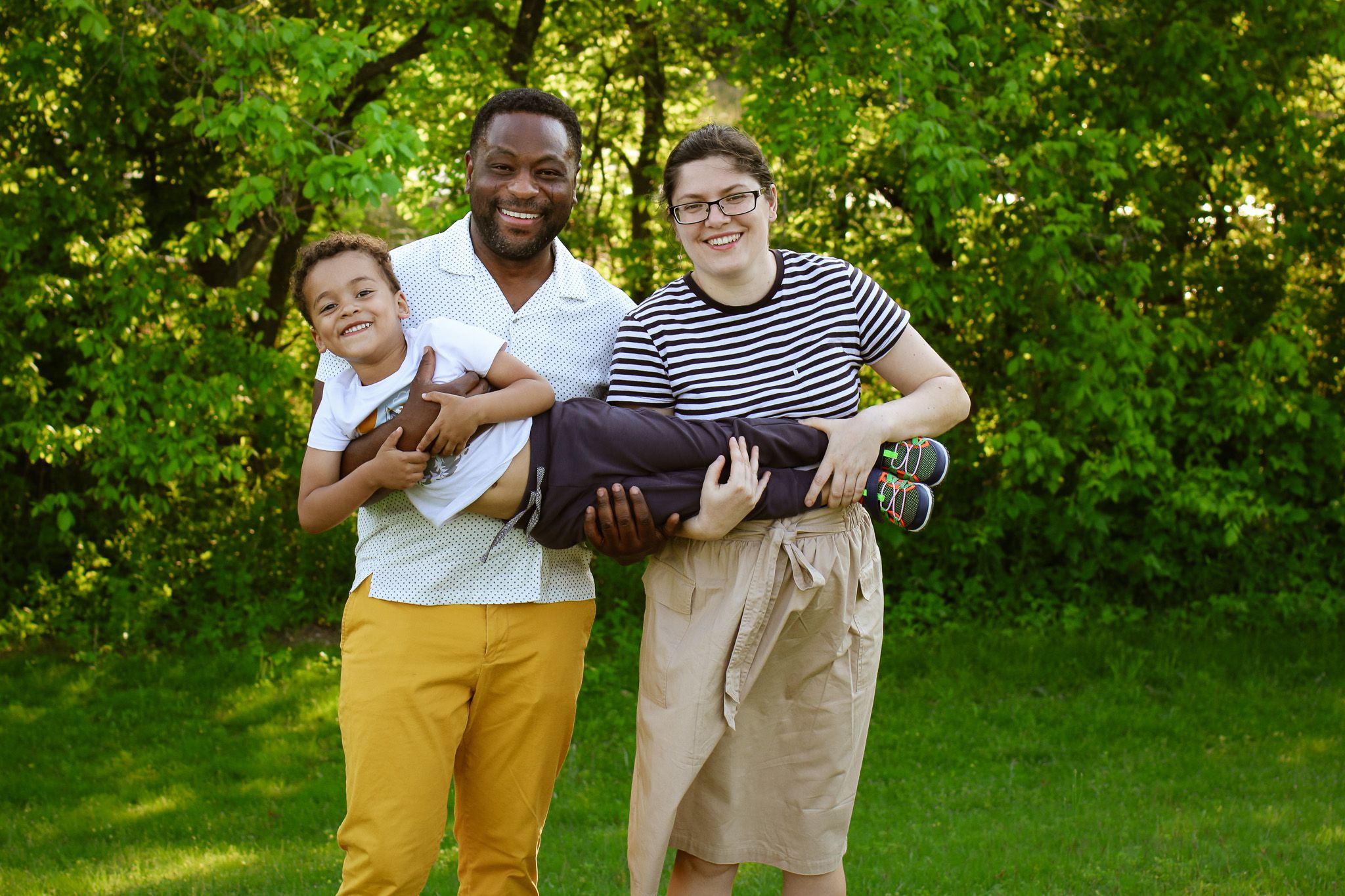 Three friends laughing while carrying their friend horizontally in a sunny garden setting.