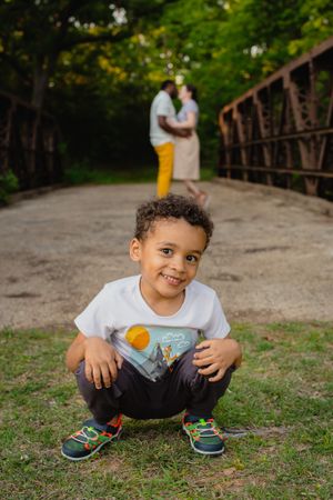 A child squats on a bridge path wearing colorful sneakers while two figures stand in soft focus in the background.