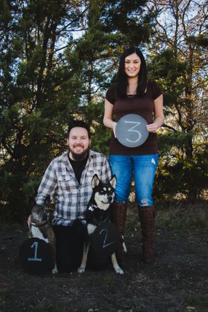 A couple with their husky dog holding numbered signs in a forest setting.
