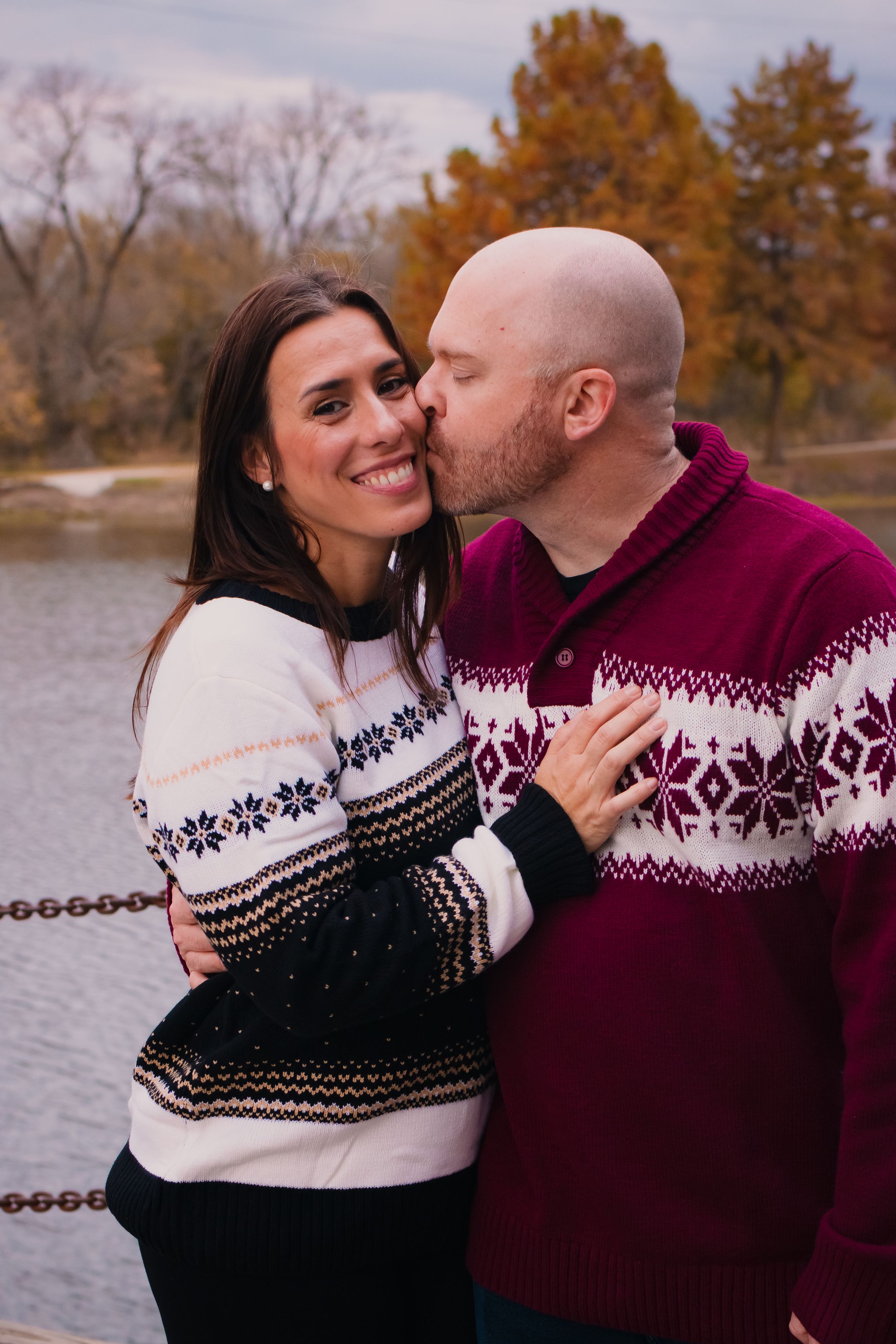 A couple wearing matching winter sweaters embraces by a lake in autumn.