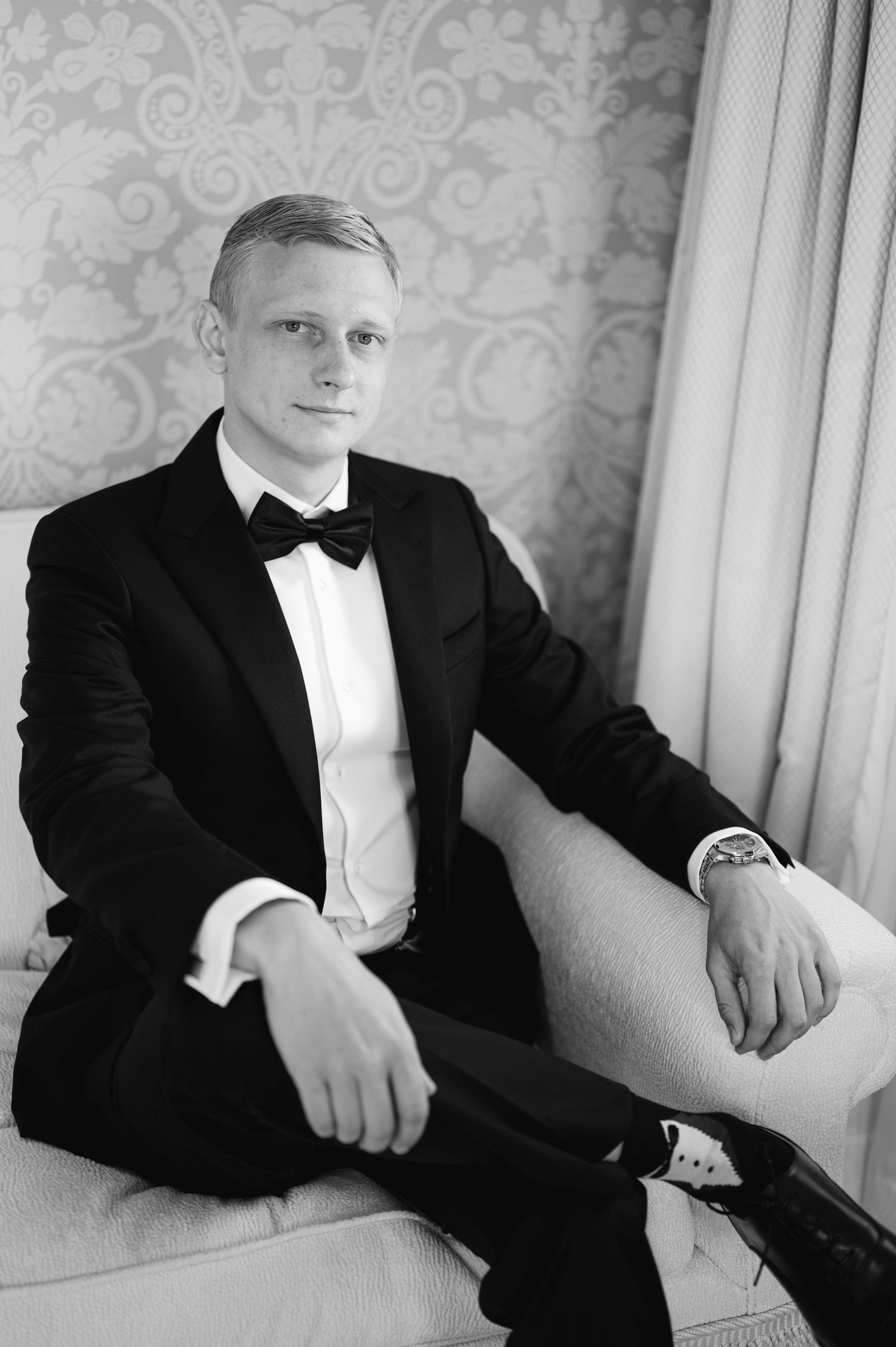 A person in a classic black tuxedo and bow tie sits in an elegant setting.
