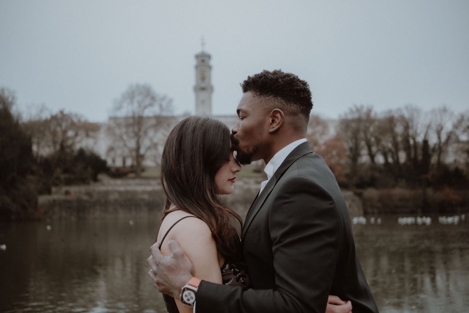 Intimate couple shares romantic moment by lake with lighthouse in background.