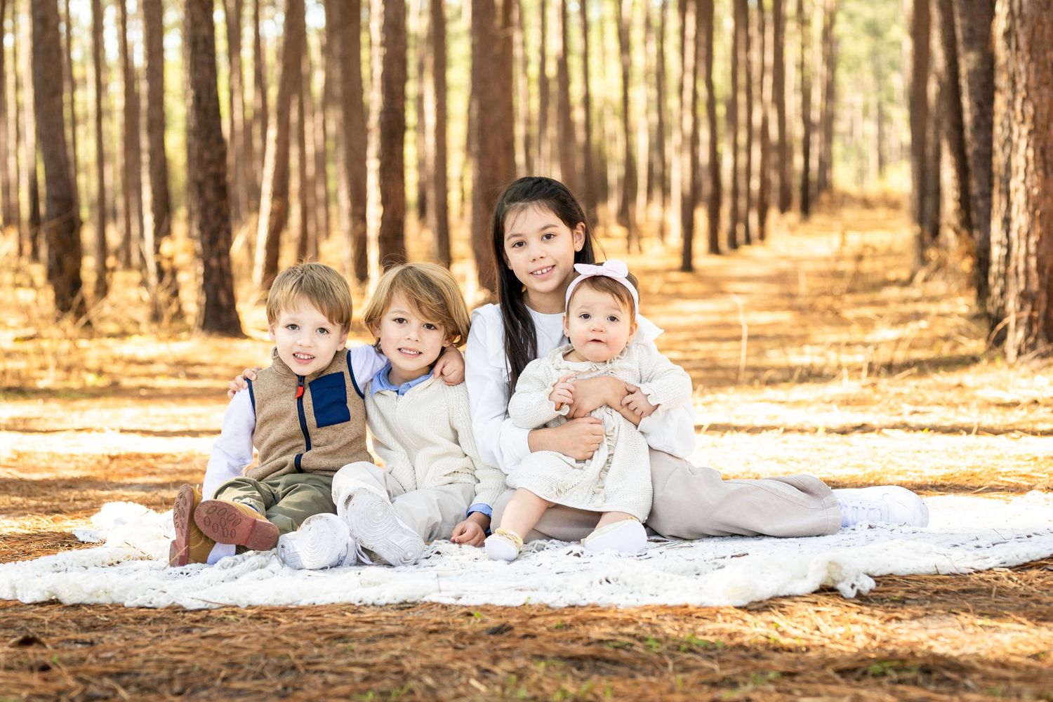 Four young children sit together on a white blanket in a sunlit pine forest.