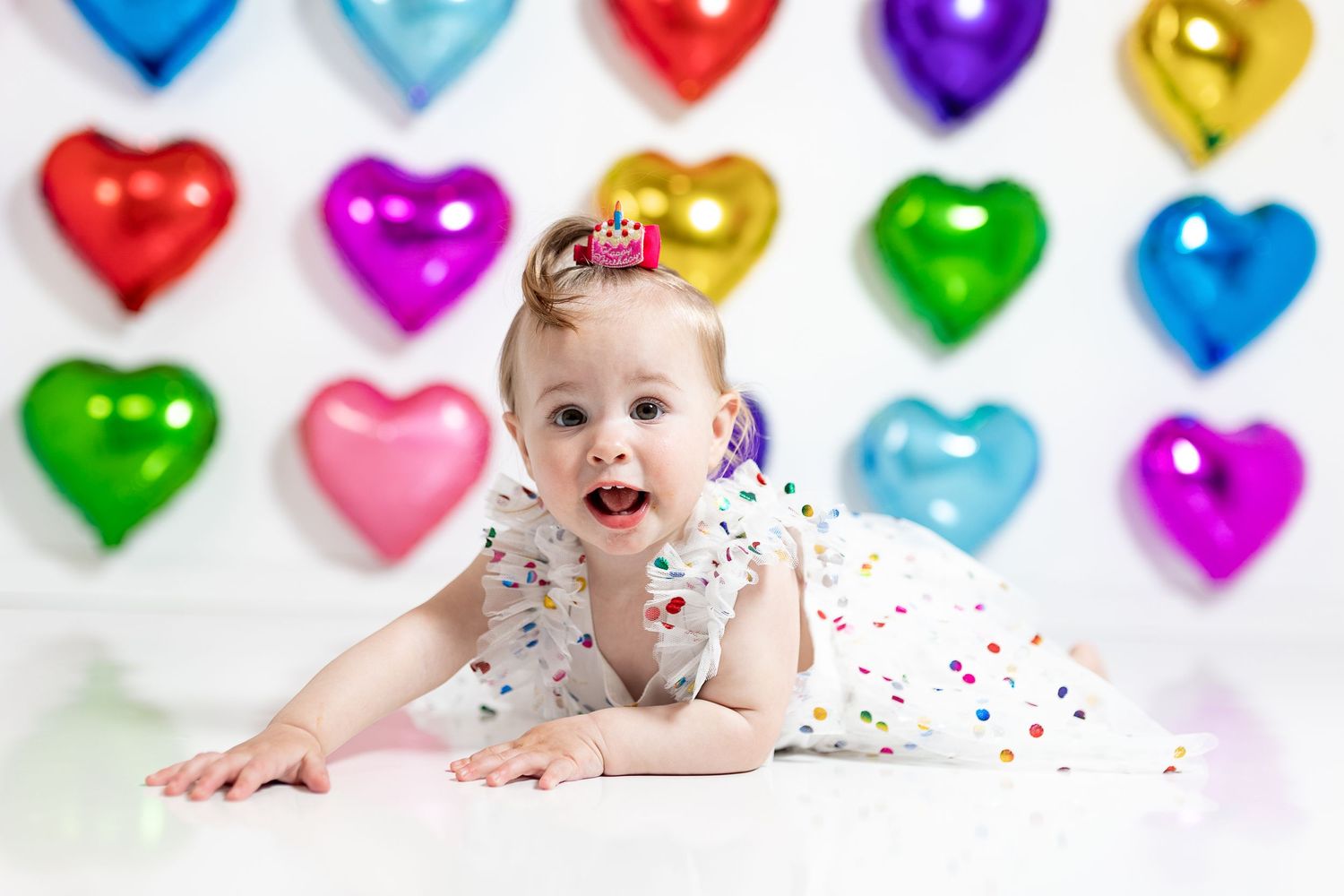 Baby in polka dot dress lies on white floor with colorful heart balloons in background.
