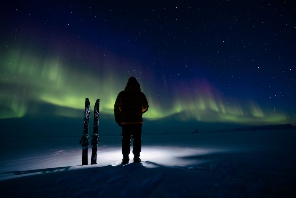 A person stands beside a splitboard in greenland looking at the aurora borealis northern lights