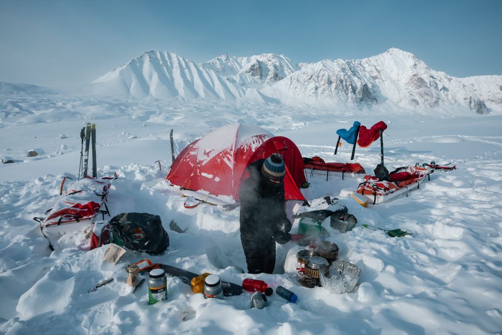 Ski tourer at a winter base camp with red tent and gear spread around the image with Stauning Alps in the background