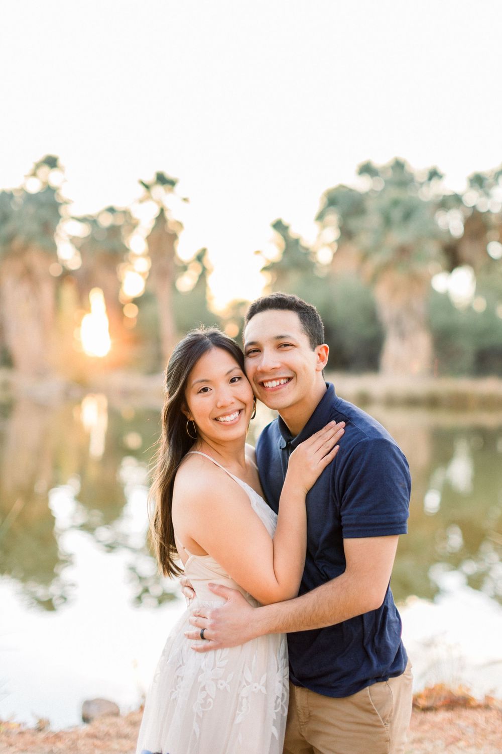 Romantic couple embraces by a tranquil lake at sunset, surrounded by palm trees.
