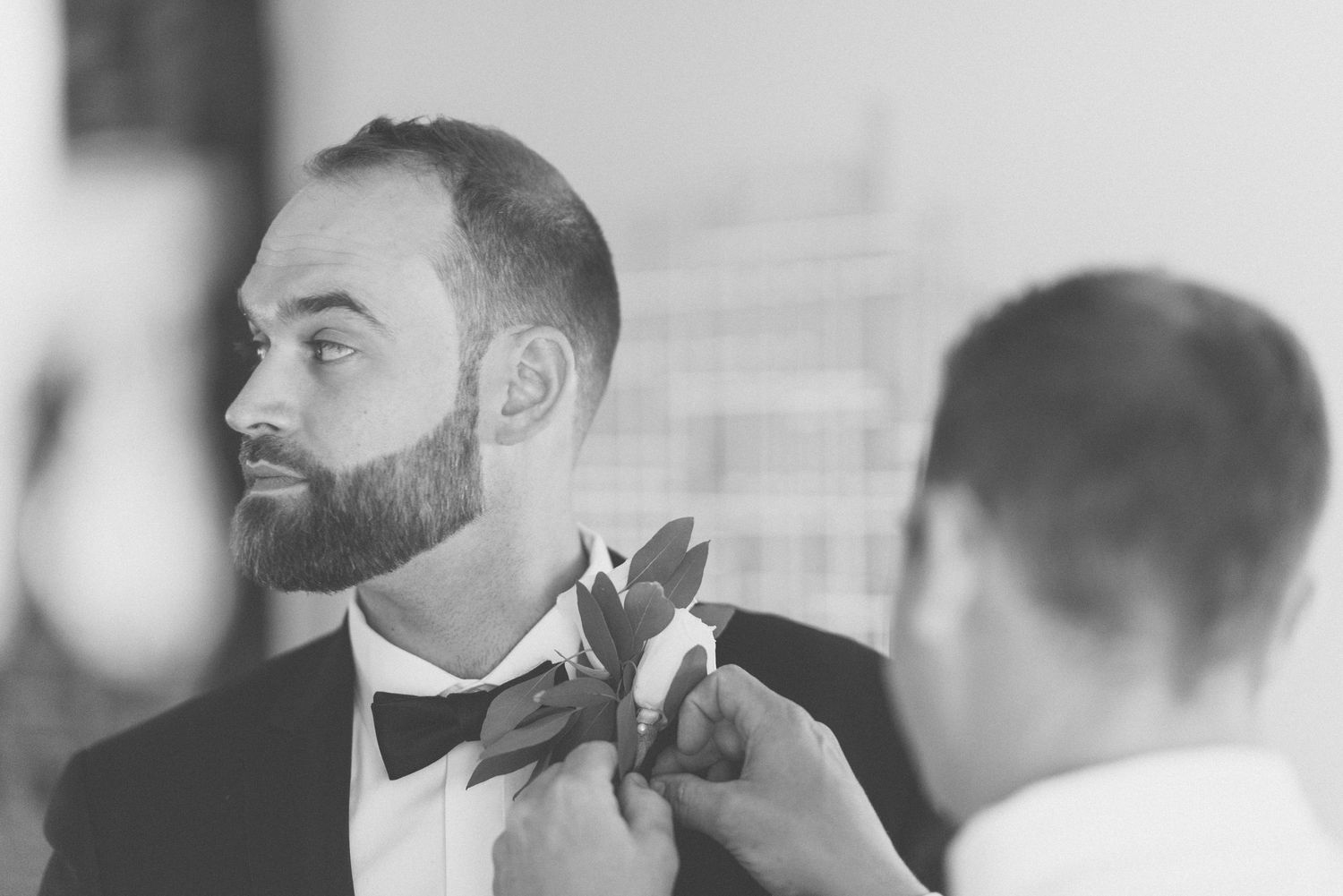 Profile view of groom in tuxedo getting boutonniere pinned on.