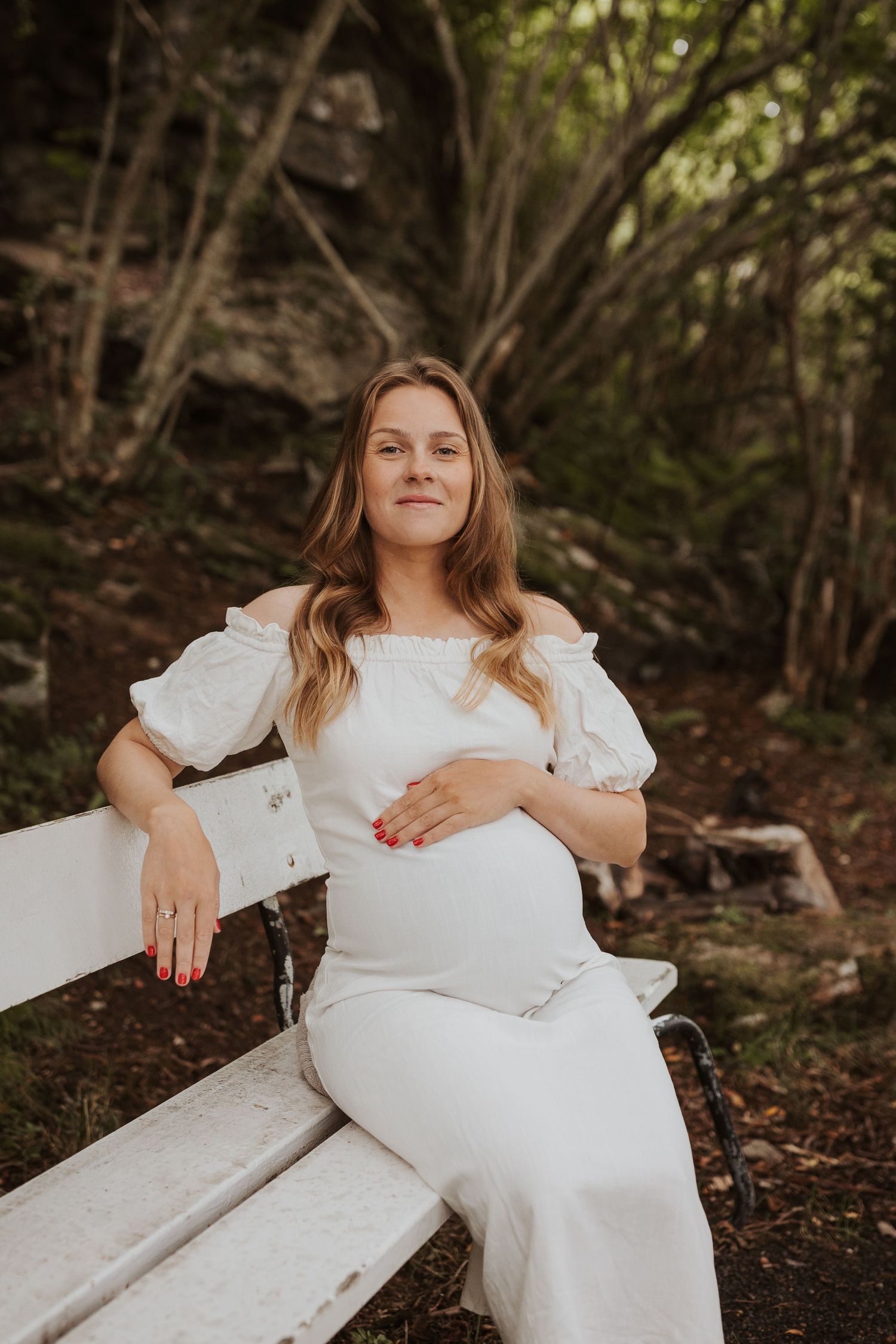 A maternity portrait of a woman in a white dress sitting on a bench in nature.