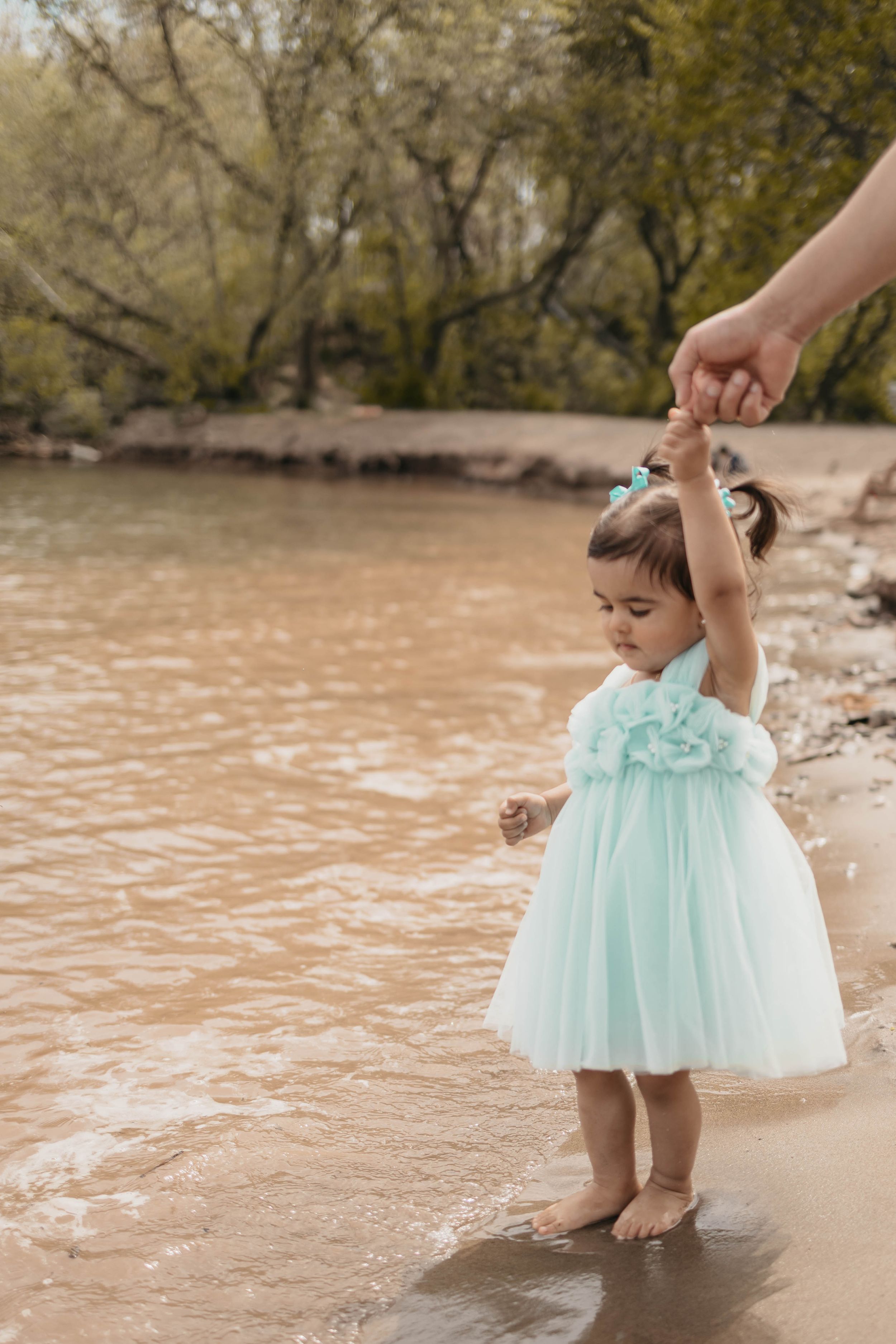 A small child in a mint tulle dress holds hands with someone while standing by the water's edge.