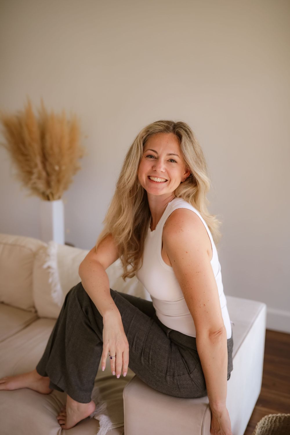Woman in white tank top and dark pants sits casually on light colored couch with pampas grass decor in background.