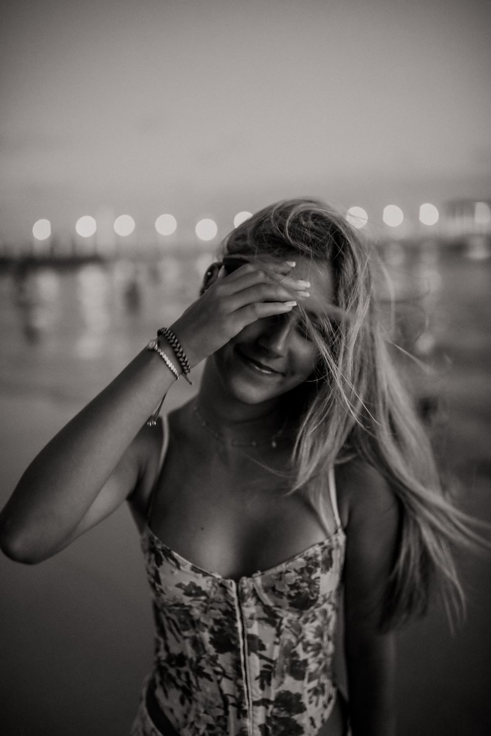 Black and white photo of someone in a floral dress laughing with hair blowing in wind at night with bokeh lights behind.